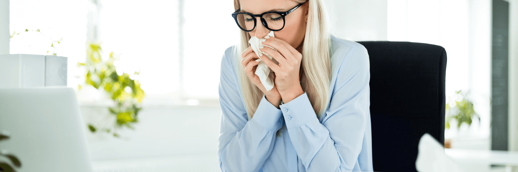 Blonde business woman wearing glasses sitting at office desk blowing nose
