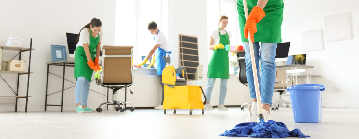 close up of woman mopping floor with apron and gloves, background is 3 members of the janitorial team cleaning office