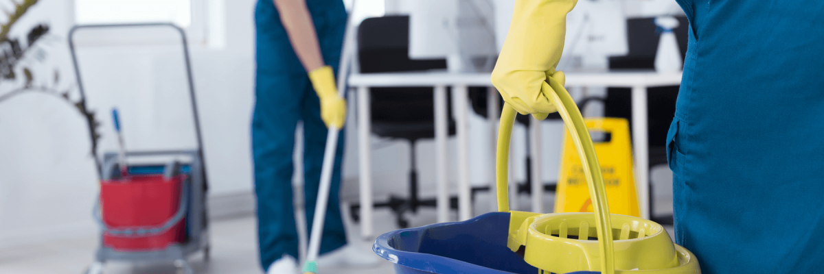 Cleaning crew in office. Individual wearing a glove holding a bucket in focus. Individual mopping in background blurred. 